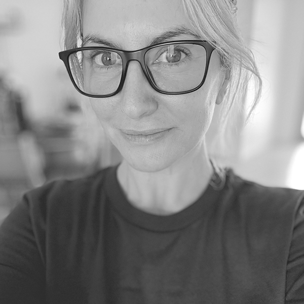 ortrait of a woman wearing glasses and a dark shirt, smiling slightly, in a softly blurred indoor setting.